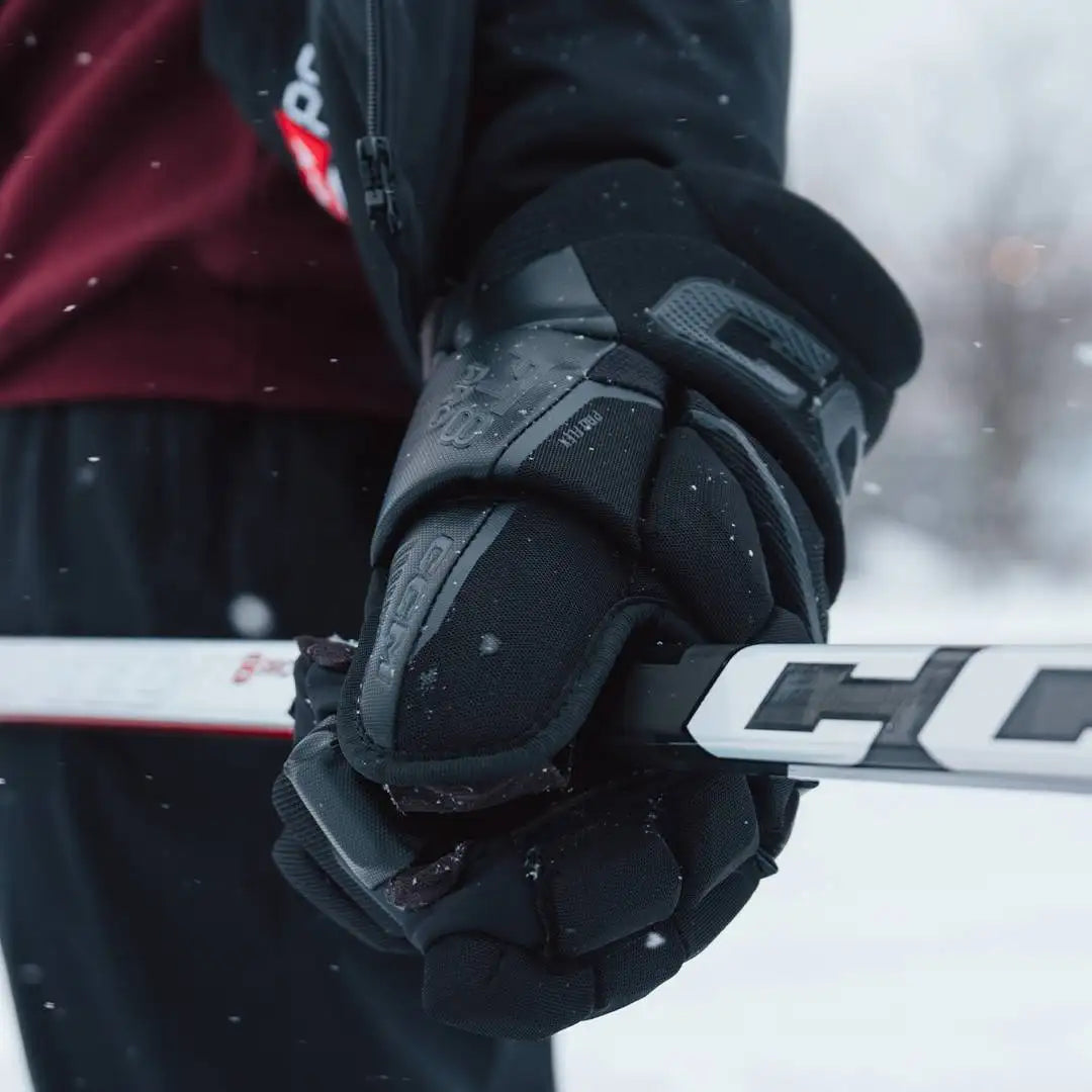 Black ccm hockey glove with gray reinforced padding and textured grip, worn on a hand gripping a white stick.